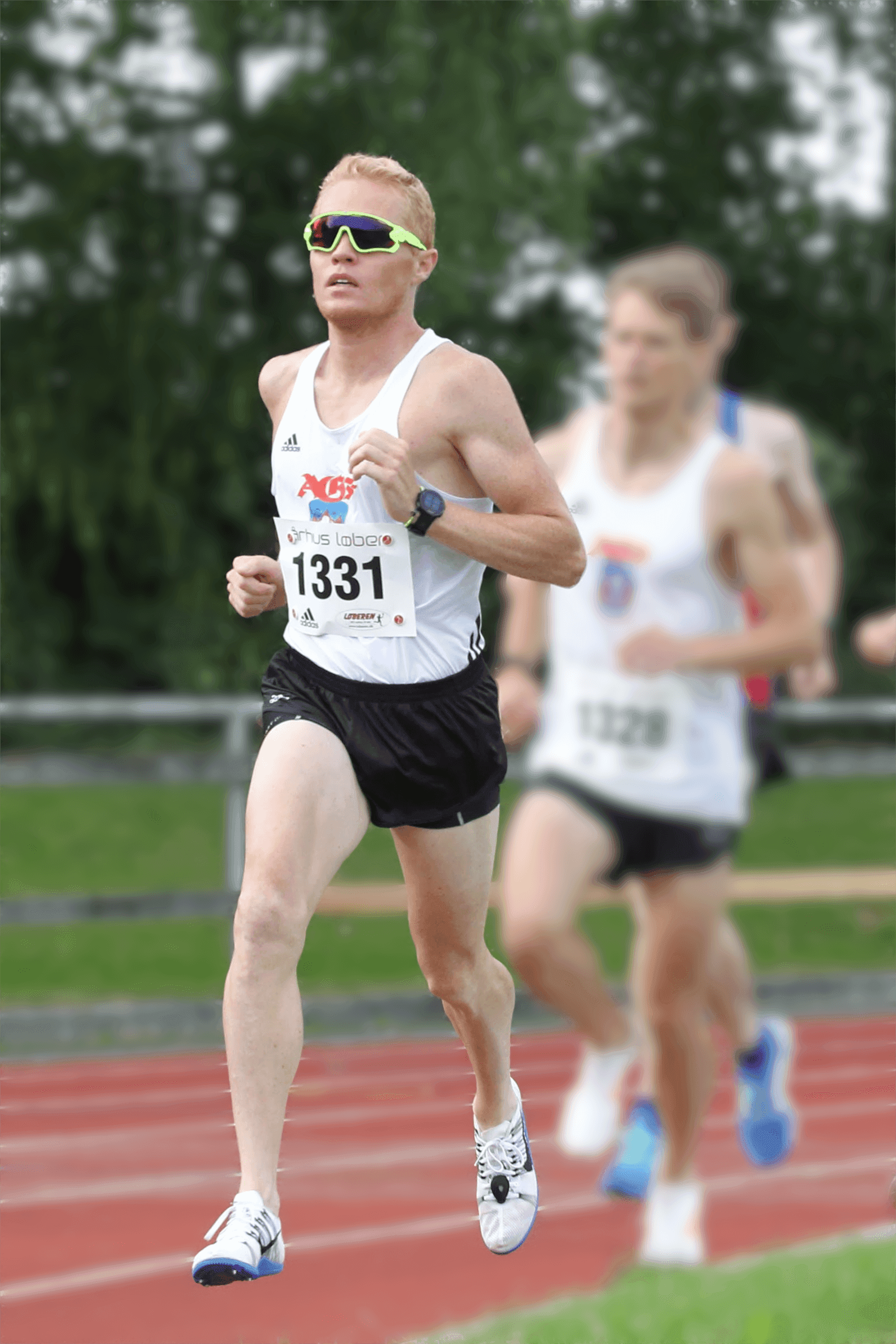 Løber i hvid tanktop og sorte shorts løber på en løbebane, iført grønne solbriller og et ur. Løbere i baggrunden er slørede. Beslutsomt udtryk.