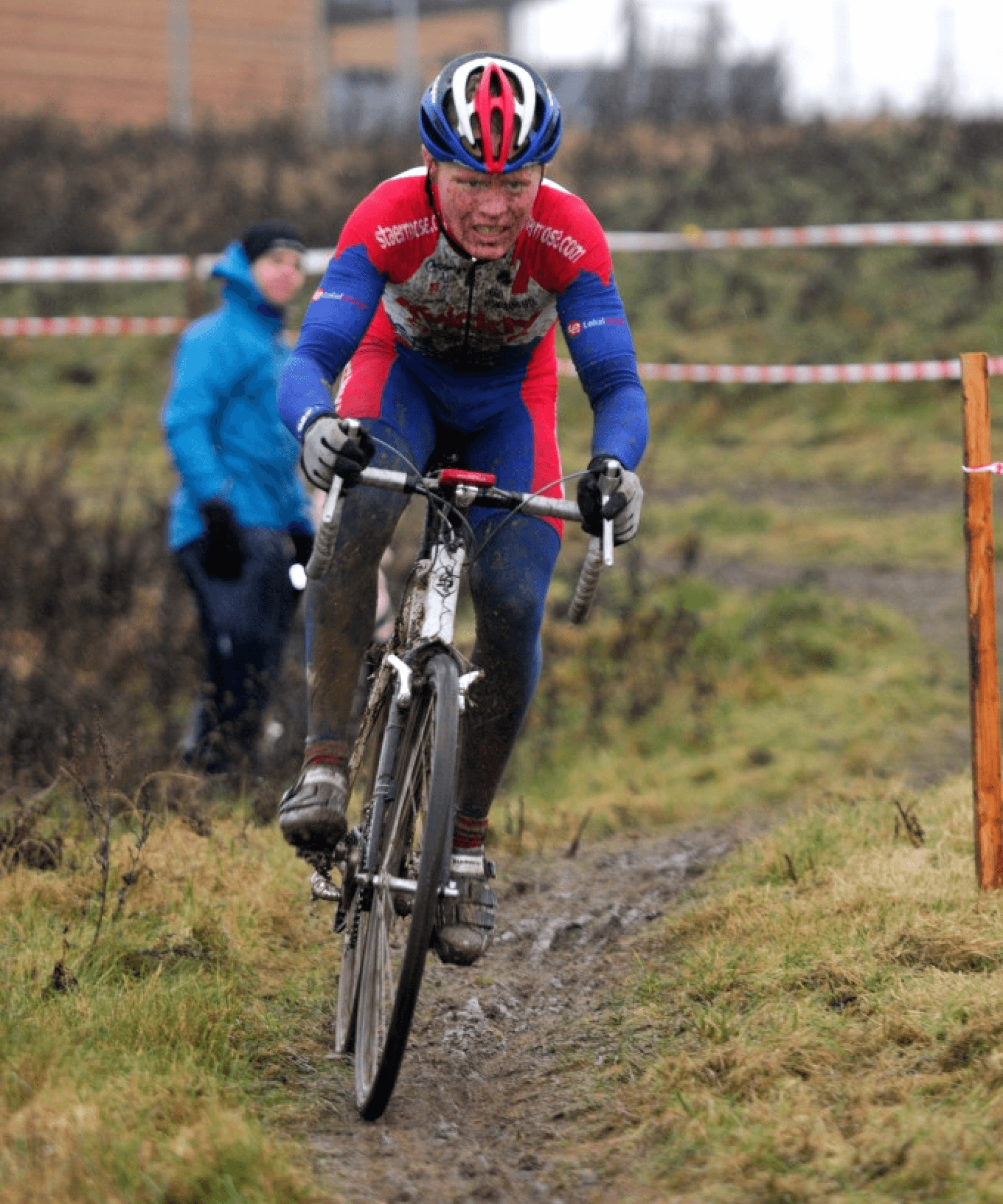 Cyclo-cross bikerider in a muddy cyclocross race, wearing a red and blue kit and helmet, rides determinedly on a grassy, wet trail. Spectator in the background in a blue jacket.