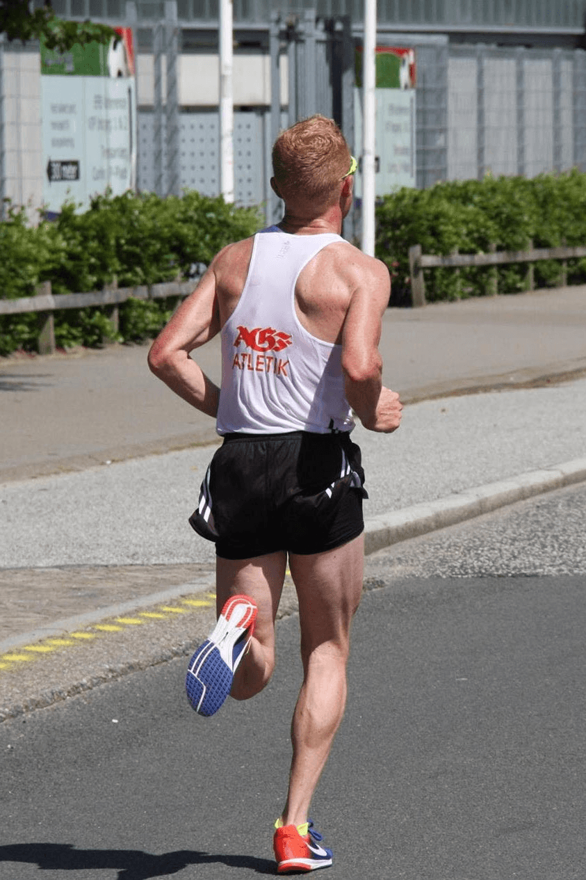 Backshot of runner in a white tank top and black shorts running on a sunny street. The text "AGF ATLETIK" is on the shirt, and greenery lines the road. Energetic tone.