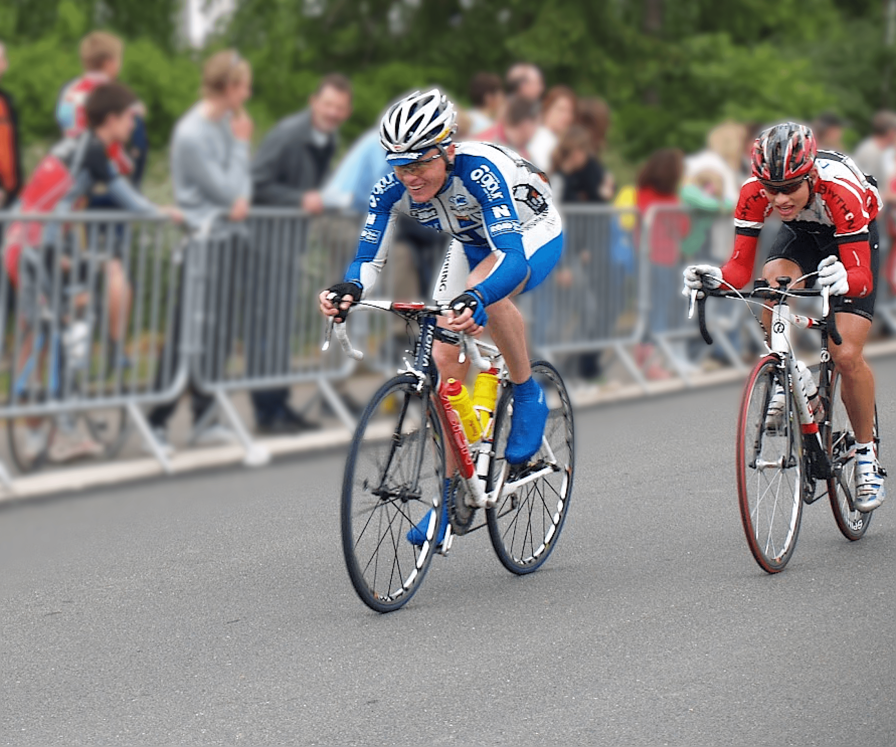 To cykelryttere i et cykelløb, fokuserede og beslutsomme, mod en baggrund af slørede tilskuere og sikkerhedsbarrierer, der formidler hastighed og konkurrenceintensitet.