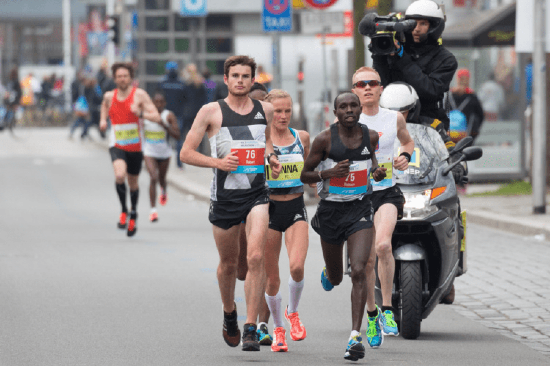 Runners compete closely in a city marathon, with bib numbers visible. A cameraman on a motorcycle captures the action. The atmosphere is intense.
