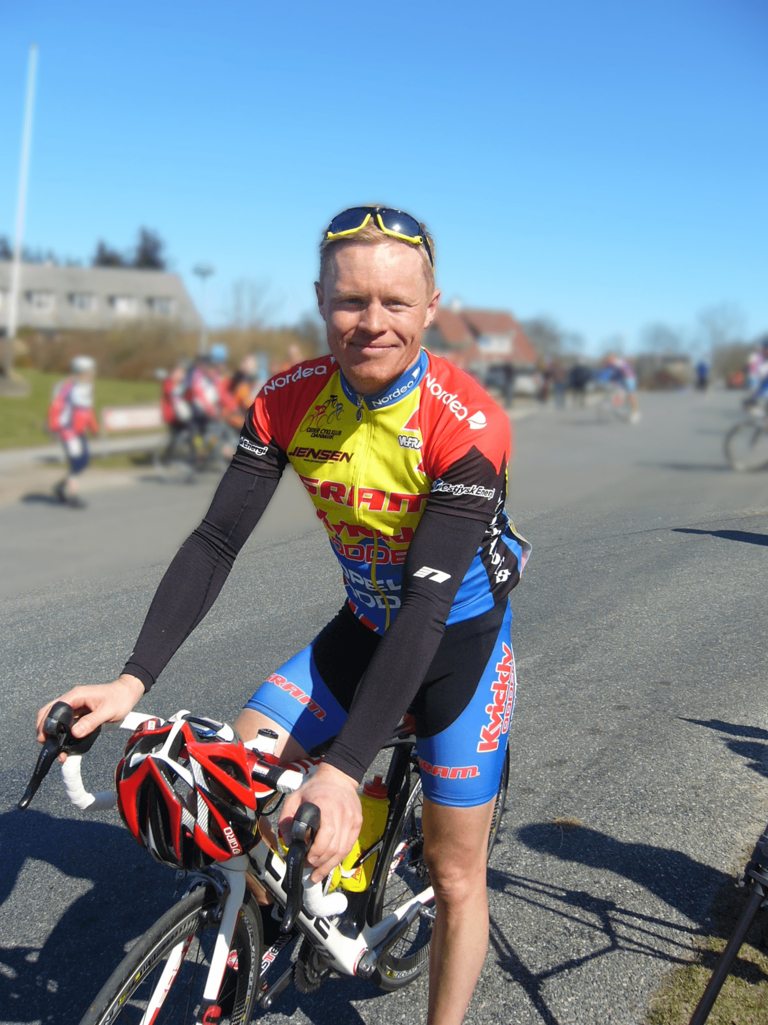 Road cyclist in a colorful racing outfit poses with his bike on a sunny day, sporting sunglasses and a helmet. Other cyclists in the background on a road.